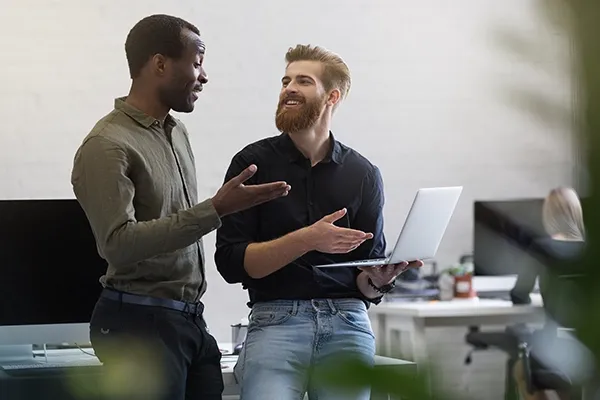 Two men standing up talking to each other with a laptop