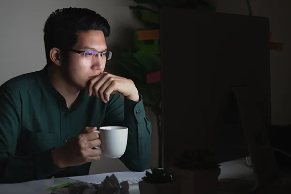 Man with glasses holding mug in right hand while looking at monitor