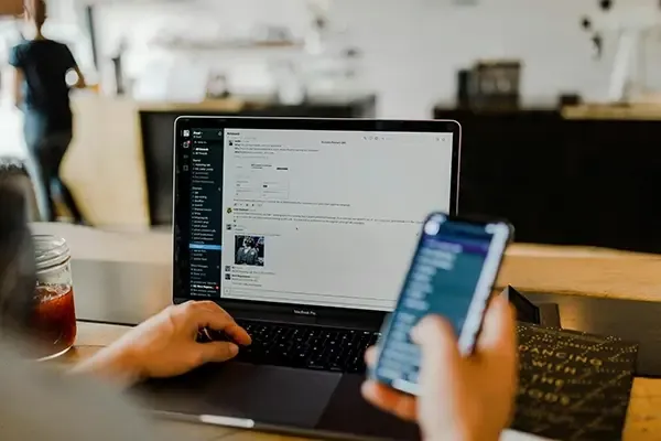 Person holding mobile phone in right hand while typing on laptop with left