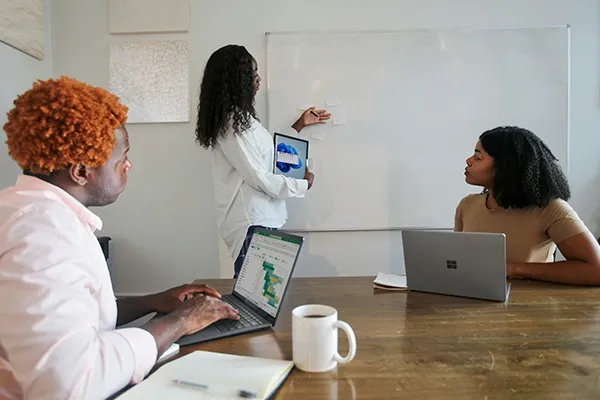 Two people sitting down with laptops looking at whiteboard